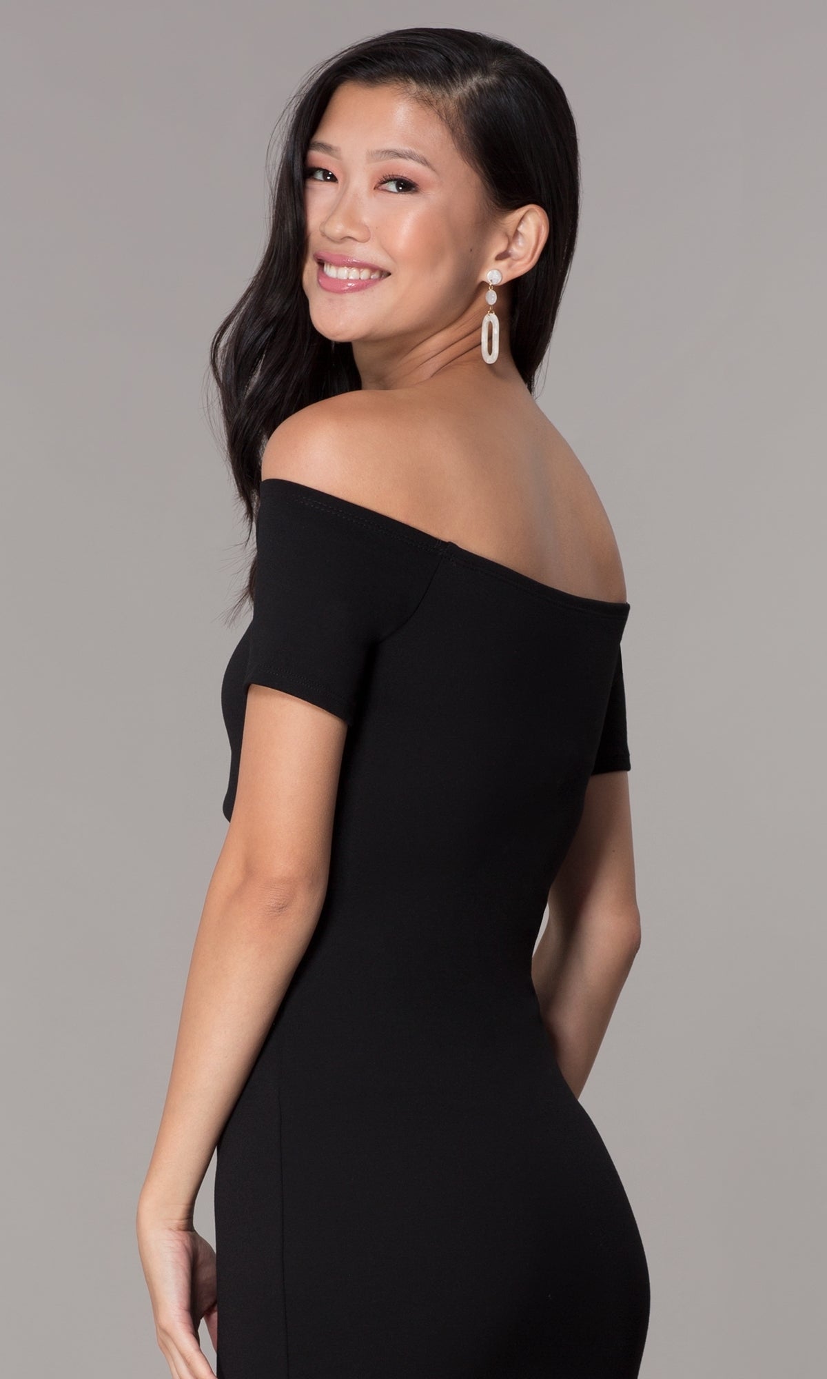 A woman wearing the Off-the-Shoulder Short Cut-Out Party Dress 3076 smiles over her shoulder, her long dark hair and dangling earrings highlighted against a plain background.